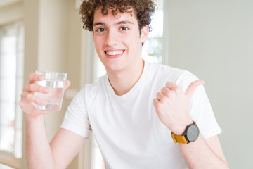 Young man drinking a glass of water at home pointing and showing with thumb up to the side with happy face smiling