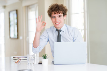 Fototapeta premium Young business man working with computer laptop at the office smiling positive doing ok sign with hand and fingers. Successful expression.