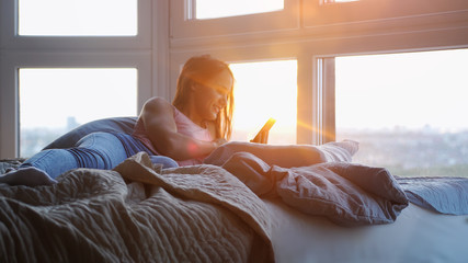 Young happy woman using smartphone on the bed during sunset
