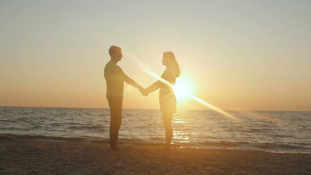 romantic young couple on the beach, man spinning around his girlfriend at sunset holding arms at beautiful seaside. Having fun together at vacation. Slow motion