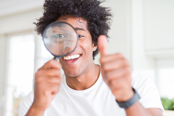 African American man looking through magnifying glass happy with big smile doing ok sign, thumb up with fingers, excellent sign - obrazy, fototapety, plakaty