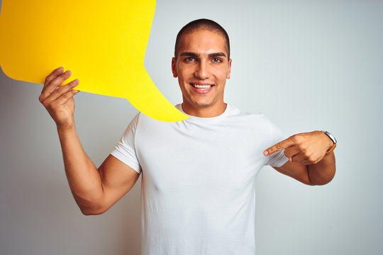 Young man holding yellow speech bubble over white isolated background with surprise face pointing finger to himself