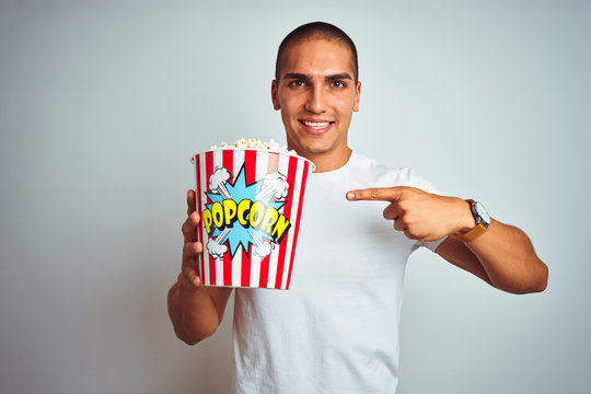 Young handsome man eating popcorn over white isolated background very happy pointing with hand and finger