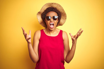 African american woman wearing summer hat and sunglasses over yellow isolated background crazy and mad shouting and yelling with aggressive expression and arms raised. Frustration concept.