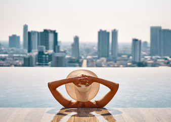 Back view of young woman in hat relaxing in swimming pool on the roof top of hotel and enjoy cityscape - obrazy, fototapety, plakaty