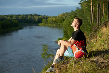 Caucasian young man with red backpack sitting with closed eyes on the brink above forest river