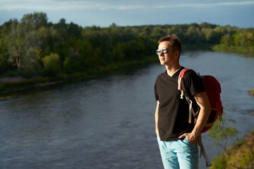 Caucasian young man with red travel backpack standing on the brink above forest river in summer and looking faraway