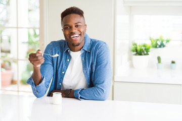 African american man eating healthy natural yogurt with a spoon with a happy face standing and...