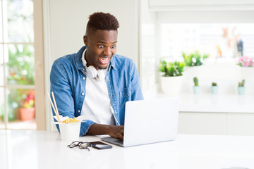 Fototapeta premium Handsome young african business man eating delivery asian food and working using computer, enjoying noodles smiling