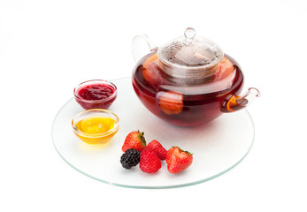 The glass teapot full of hot herbal tea with aт strawberries staying on a glass opaque tray with saucers, with honey, jam and and berries lying nearby.