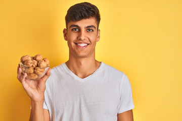 Young indian man holding bowl with walnuts standing over isolated yellow background with a happy face standing and smiling with a confident smile showing teeth