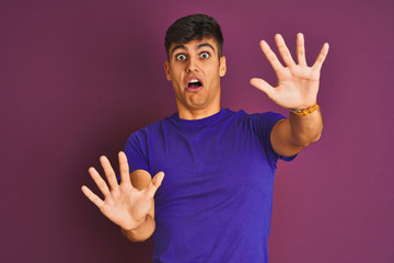 Young indian man wearing t-shirt standing over isolated purple background afraid and terrified with fear expression stop gesture with hands, shouting in shock. Panic concept.