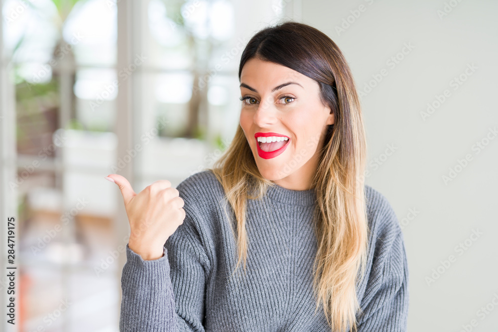 Poster Young beautiful woman wearing winter sweater at home smiling with happy face looking and pointing to the side with thumb up.