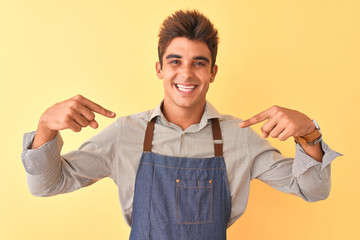 Young handsome employee man wearing apron standing over isolated yellow background looking confident with smile on face, pointing oneself with fingers proud and happy.