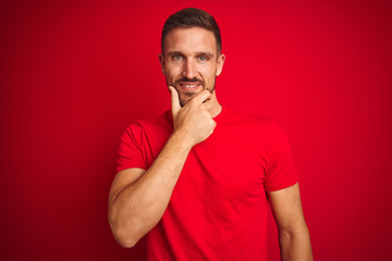 Young handsome man wearing casual t-shirt over red isolated background looking confident at the camera smiling with crossed arms and hand raised on chin. Thinking positive.
