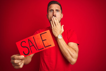 Young man holding sale advertising poster board over red isolated background cover mouth with hand shocked with shame for mistake, expression of fear, scared in silence, secret concept