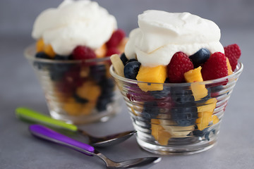 Fruit salad of banana, mango, blueberry and raspberry with whipped cream in glass glasses on a gray background.