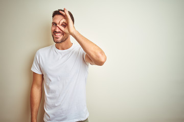 Young handsome man wearing casual white t-shirt over isolated background doing ok gesture with hand smiling, eye looking through fingers with happy face.