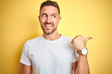 Young handsome man wearing casual white t-shirt over yellow isolated background smiling with happy face looking and pointing to the side with thumb up.