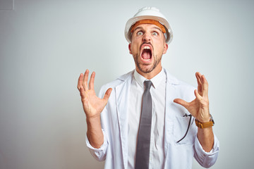 Young handsome engineer man wearing safety helmet over isolated background crazy and mad shouting and yelling with aggressive expression and arms raised. Frustration concept.