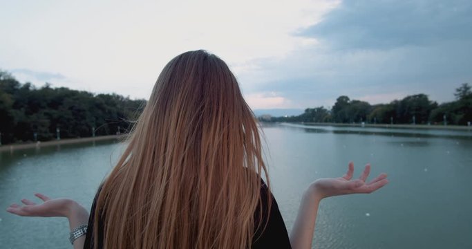 Young woman makes hand gesture that means I don't know, standing on river bridge