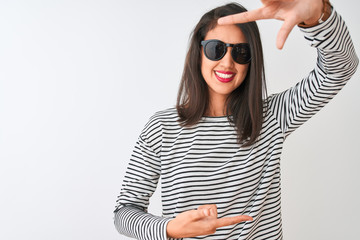 Chinese woman wearing striped t-shirt and sunglasses standing over isolated white background smiling making frame with hands and fingers with happy face. Creativity and photography concept.