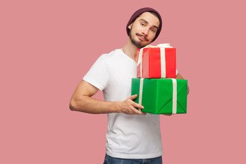 Portrait of handsome bearded young hipster man in white shirt and casual hat standing and holding two present box, looking at camera. Indoor, isolated, studio shot, pink background