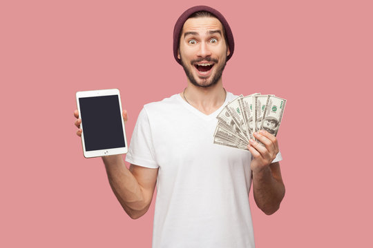 Portrait Of Suprised Handsome Bearded Young Hipster Man In White Shirt And Casual Hat Standing, Holding Empty Screen Ipad And Green Present Box. Indoor, Isolated, Studio Shot, Pink Background