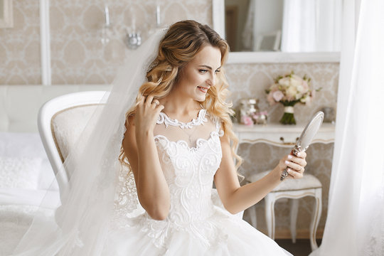 Beautiful bride in luxury wedding dress keeping the mirror and looking on her reflexion and posing in interior