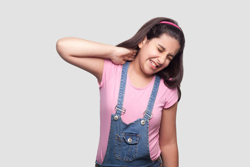 Neck pain. Portrait of sad brunette young girl in pink t-shirt and blue overalls standing and holding her painful neck and feeling bad. indoor studio shot, isolated on light gray background.
