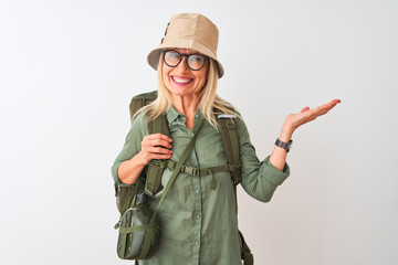 Middle age hiker woman wearing backpack hat canteen glasses over isolated white background smiling cheerful presenting and pointing with palm of hand looking at the camera.