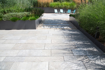 Moderne Garten- und Terrassengestaltung im Materialmix: Terrasse und Gehweg aus Steinplatten umgeben von Schotter und Metall Pflanzgefäßen mit Grünpflanzen