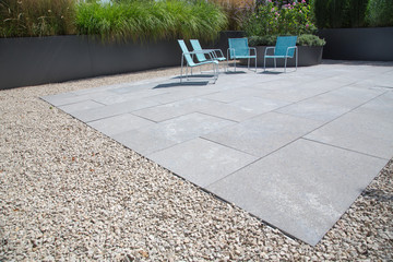 Moderne Garten- und Terrassengestaltung im Materialmix: Terrasse aus Steinplatten umgeben von Schotter und Metall Pflanzgefäßen mit Grünpflanzen
