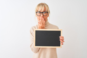 Middle age woman wearing glasses holding blackboard over isolated white background cover mouth with hand shocked with shame for mistake, expression of fear, scared in silence, secret concept