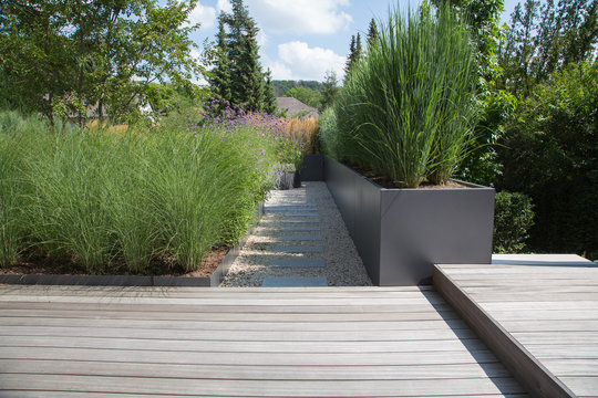 Moderne Garten- und Terrassengestaltung im Materialmix: Terrassen aus Holz und Gehweg aus Steinplatten umgeben von Schotter und Metall Pflanzgefäßen mit Grünpflanzen 