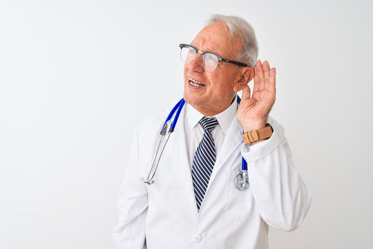 Senior grey-haired doctor man wearing stethoscope standing over isolated white background smiling with hand over ear listening an hearing to rumor or gossip. Deafness concept.