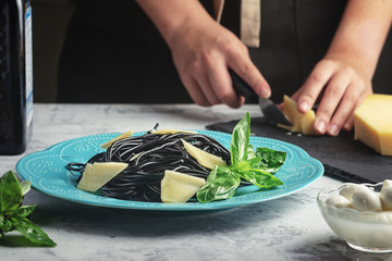 Girl cuts cheese to decorate black spaghetti dishes. Unusual cuttlefish ink pasta. A wholesome light dinner with basil.
