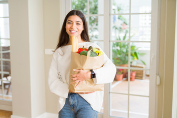 Beautiful young woman smiling holding a paper bag full of fresh groceries at home