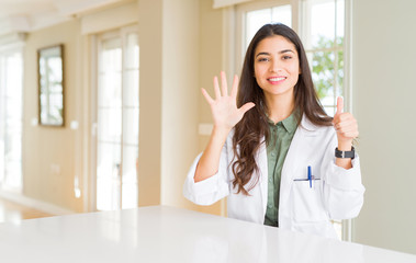 Young woman wearing medical coat at the clinic as therapist or doctor showing and pointing up with fingers number six while smiling confident and happy.