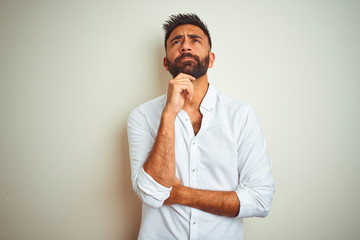 Young indian man wearing elegant shirt standing over isolated white background with hand on chin...