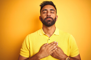 Young indian man wearing polo standing over isolated yellow background smiling with hands on chest with closed eyes and grateful gesture on face. Health concept.