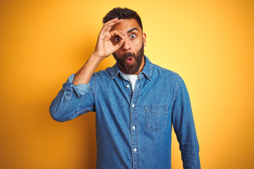 Young indian man wearing denim shirt standing over isolated yellow background doing ok gesture shocked with surprised face, eye looking through fingers. Unbelieving expression.