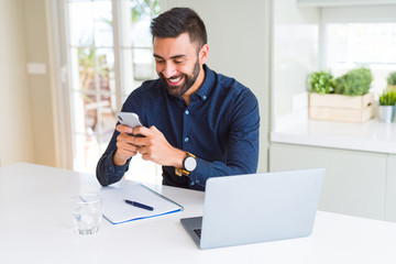 Handsome hispanic business man using smartphone and laptop at the office with a happy face standing...