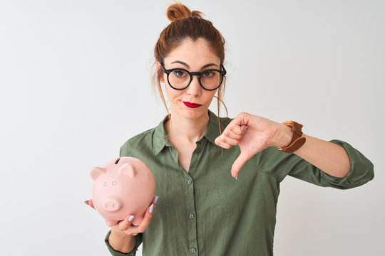 Beautiful redhead woman wearing glasses holding piggy bank over isolated white background with angry face, negative sign showing dislike with thumbs down, rejection concept