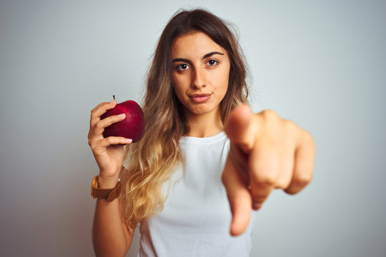 Young beautiful woman eating red apple over grey isolated background pointing with finger to the camera and to you, hand sign, positive and confident gesture from the front