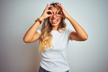 Obraz na płótnie Canvas Young beautiful woman wearing casual white t-shirt over isolated background doing ok gesture like binoculars sticking tongue out, eyes looking through fingers. Crazy expression.