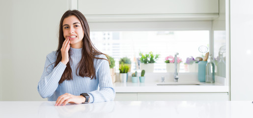 Wide angle picture of beautiful young woman sitting on white table at home touching mouth with hand with painful expression because of toothache or dental illness on teeth. Dentist concept.