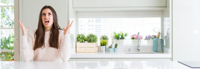 Wide angle picture of beautiful young woman sitting on white table at home crazy and mad shouting and yelling with aggressive expression and arms raised. Frustration concept.