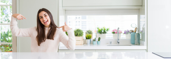 Wide angle picture of beautiful young woman sitting on white table at home looking confident with smile on face, pointing oneself with fingers proud and happy.