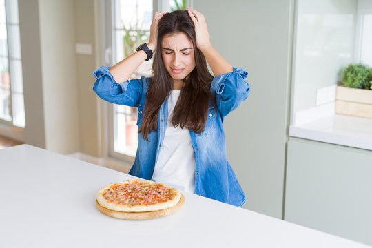 Beautiful young woman eating homemade tasty pizza at the kitchen suffering from headache desperate and stressed because pain and migraine. Hands on head.